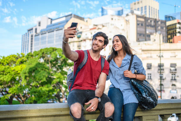 Front view close-up of relaxed young Hispanic couple sitting on balustrade and taking selfie with downtown Buenos Aires in background.